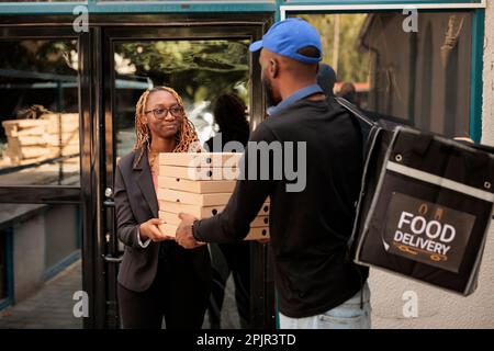 Essenslieferung ins Büro, fröhlicher Kunde mit Pizzakartonstapel, Frau erhält Bestellung vom Kurier im Freien. afroamerikanischer Angestellter nimmt Essenspakete auf, Mann liefert Mittagessen Stockfoto