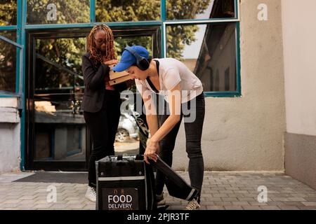 Kurier in Kopfhörern holt Pizzaschachteln aus dem Thermosack, Kunde erhält Pizzeria-Bestellung vor dem Bürogebäude im Freien. Essenslieferservice, Frau, die Essen zum Mitnehmen liefert Stockfoto