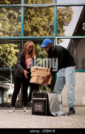 Kurier fährt an pizzakartons von afroamerikanischer Frau vorbei, die sich in der Nähe des Bürogebäudes stapeln, Pizzeria-Lieferservice. Ein Mann, der eine Bestellung aus einem Thermal-Rucksack abnimmt, ein Lieferant, der dem Gast Fastfood aus dem Café gibt Stockfoto