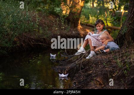 Mädchen und Junge sitzen am Ufer des Flusses und werfen das Origami-Boot aus weißem Papier ins Wasser. Eine Teenager-Schwester mit grünem Haar umarmt den kleinen Bruder. Stockfoto