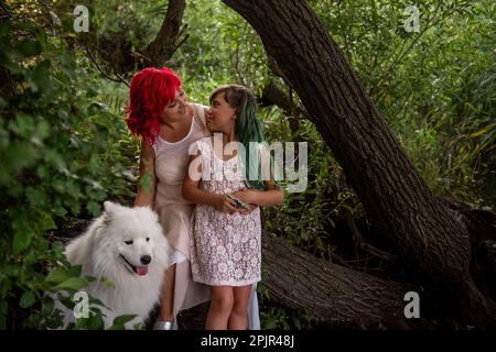 Im Grünen Wald, an einem kleinen Diversitätsstrom, umarmte Mutter mit rosa Haaren Tochter. Der weiße, flauschige samoyed bewacht die Familie. Reisen mit Haustier Stockfoto