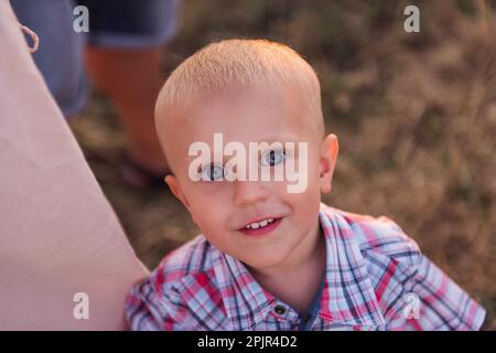 Nahaufnahme eines kleinen Jungen im karierten Hemd, umarmtes Mütterbein. Draufsicht auf ein lächelndes Kleinkind mit blauen Augen. Der Begriff des elterlichen Schutzes, t Stockfoto