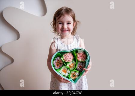 Das kleine lockige Mädchen hält eine herzförmige Box mit hausgemachten, süßen Marshmallows in den Händen. Natürliche, zuckerfreie Öko-Produkte. Essbares Geschenk, Blumenstrauß Stockfoto