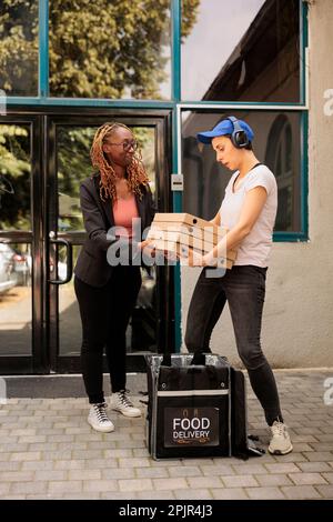 Kurierfahrer in Kopfhörern, die den Kunden Pizzastücke stapeln lassen, Frau, die Bestellungen aus dem Thermal-Rucksack vor dem Bürogebäude im Freien aufnimmt. Pizzeria-Food-Lieferservice Stockfoto