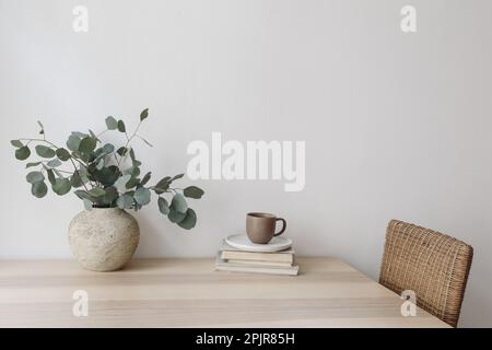 Silbergrüne Eukalyptusäste in beigefarbener strukturierter Vase. Braune Tasse Kaffee, Tee und alte Bücher auf einem Holztisch. Weißer Wandhintergrund Stockfoto