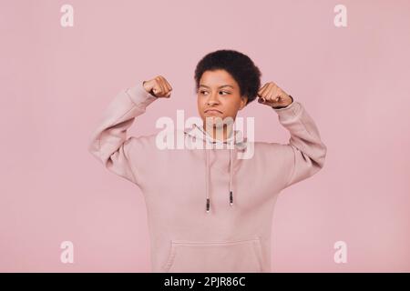 Mutige Studentin in Kapuzenpullover mit pinkfarbener Kulisse. Starke schwarze junge Frau, die ihre Hände vor pinkfarbenem Hintergrund hebt Stockfoto