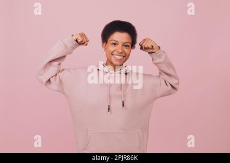 Mutige, fröhliche junge Frau in Kapuzenpullover posiert mit erhobenen Händen. Studio-Porträt eines schwarz lächelnden Mädchens im Hoodie mit pinkfarbenem Hintergrund Stockfoto