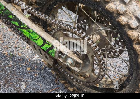 Kette und schwarzer hinterer Gummireifen, der mit getrocknetem Schlamm auf einem auf Schotterstraßen geparkten Motorrad bedeckt ist. Stockfoto