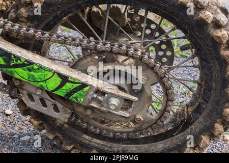 Kette und schwarzer hinterer Gummireifen, der mit getrocknetem Schlamm auf einem auf Schotterstraßen geparkten Motorrad bedeckt ist. Stockfoto