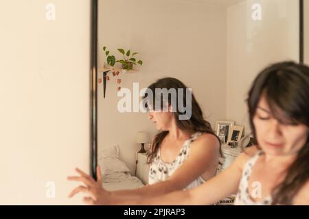 Hispanische junge Erwachsene Frau, die sich ein Kleid aus dem Kleiderschrank im Schlafzimmer zu Hause aussucht, über einen Spiegel reflektiert, trauriges, deprimierendes symbolisches Bild Stockfoto