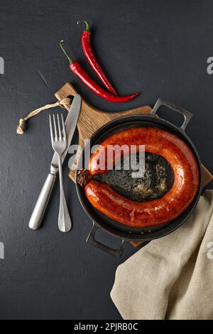 Gegrillte Schweinewurst in einem Ring in einer gusseisernen Pfanne, heiße Paprika, Besteck und eine Leinenserviette auf einem dunklen Steintisch. Draufsicht mit Kopierbereich Stockfoto