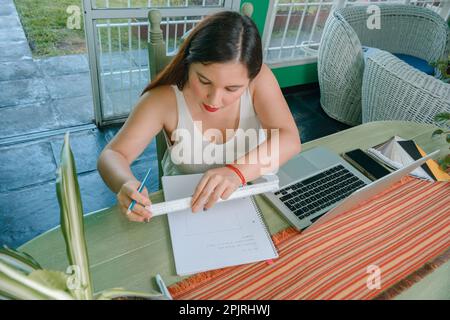 Junge Latina-Frau argentinischer Herkunft, Innenarchitekt, die zu Hause mit ihrem Computer und mit einem Notizbuch arbeitet, das eine Spur auf der Seite macht Stockfoto