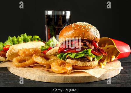 Leckere Burger, Pommes Frites, gebratene Zwiebelringe, Hot Dog und erfrischendes Getränk auf einem schwarzen Holztisch. Fastfood Stockfoto