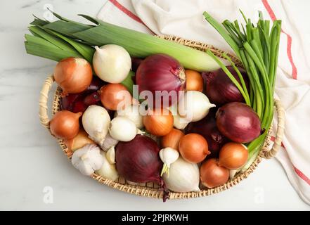 Weidenschale mit frischen Zwiebelknollen, Lauch und Knoblauch auf weißem Marmortisch, flach liegend Stockfoto