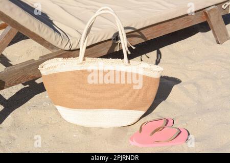 Strohbeutel und Flip Flops in der Nähe einer Holzliege am Sandstrand. Sommeraccessoires Stockfoto