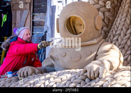 PRODUKTION - 03. April 2023, Mecklenburg-Vorpommern, Warnemünde: Sandkünstler Andrius Petkus aus Litauen arbeitet mit einem Taucher an einer großen Sandskulptur. Sechs Künstler aus fünf Ländern kreieren die Figuren für die 13. Sand World aus 300 Tonnen Spezialsand direkt am Meereskanal in Warnemünde. Unter dem Motto „Metamorphose - Blown by the (Baltic) Wind“ werden bis zum 06. April 2023 verschiedene Motive rund um das Thema Meer geschaffen. Die Show wird bis zum 29. Oktober 2023 ausgestellt. Foto: Jens Büttner/dpa Stockfoto