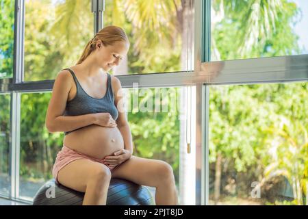 Schwangere Frau, die zu Hause auf Sportball trainiert. Schwangere Frau, die Entspannungsübungen mit einem Fitness-Pilates-Ball macht. Vor dem Hintergrund des Fensters Stockfoto
