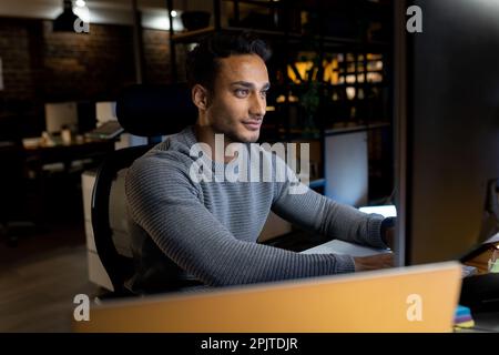 Glücklicher birassistischer Geschäftsmann, der am Schreibtisch sitzt, Computer benutzt, bis spät in die Nacht im Büro arbeitet Stockfoto