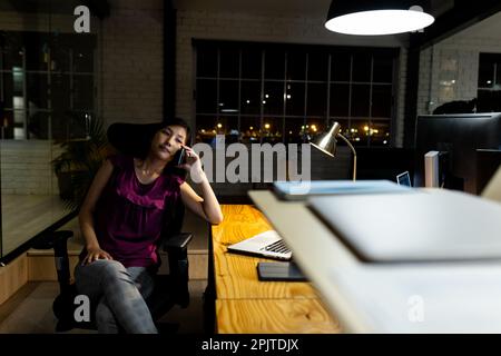 Asiatische Geschäftsfrau sitzt am Schreibtisch, telefoniert über ein Smartphone, arbeitet bis spät in die Nacht im Büro Stockfoto