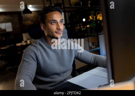 Glücklicher birassistischer Geschäftsmann, der am Schreibtisch sitzt, Computer benutzt, bis spät in die Nacht im Büro arbeitet Stockfoto