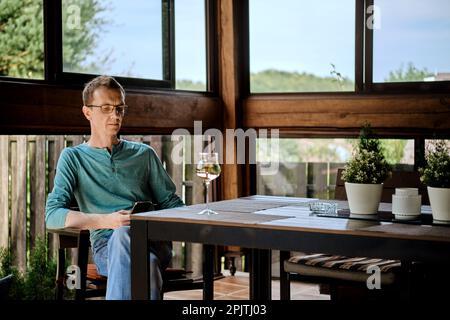 Ein Mann mittleren Alters sitzt auf der Veranda des Landhauses mit einem Glas Wein und benutzt ein Smartphone Stockfoto