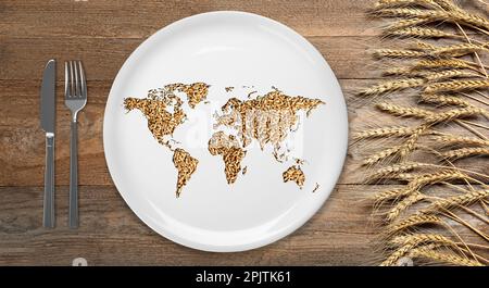 Das Konzept der globalen Nahrungsmittelkrise. Weltkarte aus Weizenkörnern in Teller, Besteck und Stacheln auf Holztisch, flach liegend Stockfoto