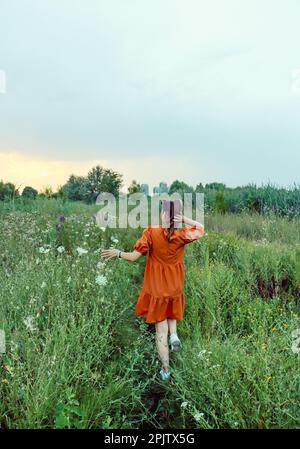 Ein Mädchen in einem orangefarbenen Kleid reist allein durch das Feld der Frühlingsblumen und bewundert die Farben des Sonnenuntergangs Stockfoto