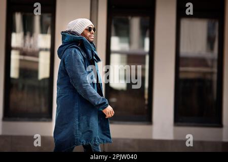 Porträt eines gutaussehenden afroamerikanischen Mannes, der in der Stadt spaziert, einen stylischen Parka-Mantel, einen Strickmütze und eine Sonnenbrille trägt. Street Style Mode Männchen Mo Stockfoto