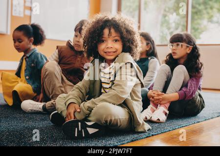 Der Junge sitzt in einer Grundschule und schaut in die Kamera. Ein multiethnisches Kind besucht eine gemeinsame Grundschule mit einer Gruppe multiethnischer Kinder Stockfoto