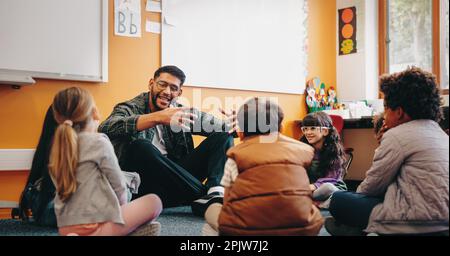 Männlicher Erzieher, der mit seinen Schülern in einem Klassenzimmer spricht. Ein Mann, der Grundschulkinder unterrichtet, sitzt mit ihnen auf dem Boden und gibt ein Wort Stockfoto