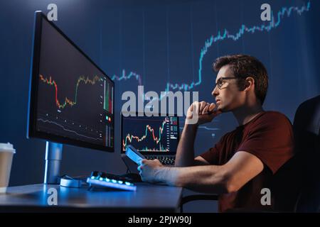 Seitenansicht eines konzentrierten Krypto-Händlers, der vor Computern sitzt, professionelle Analyse von Kerzenständerdiagrammen macht, Strategie erstellt, Stift und Notizblock hält, auf den Monitor schaut, das Kinn berührt Stockfoto