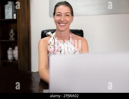 Eine reife Geschäftsfrau sitzt an ihrem Schreibtisch und lächelt vor Freude, während sie an ihrem Laptop im Büro arbeitet. Stockfoto