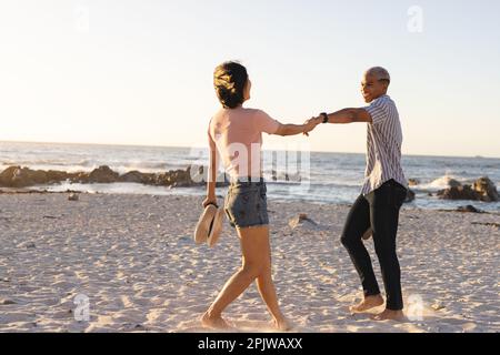 Glückliches, vielfältiges Paar, das Händchen hielt und am Strand spazierte Stockfoto