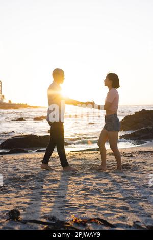 Ein glückliches, vielfältiges Paar, das sich am Strand an den Händen hält und gemeinsam tanzt Stockfoto