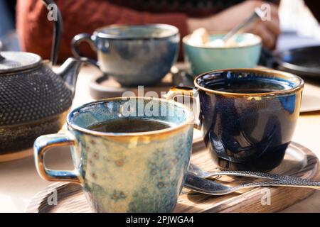 Tee zubereiten und servieren. Teekanne, grüner Tee, blauer Keramikbecher auf einem hölzernen Café-Tisch. Zwei handgemachte Töpferbecher am frühen Morgen. Trendiger Autor Stockfoto