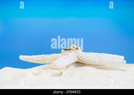 Goldene Hochzeitsringe auf weißen Seesternen am Sandstrand. Marineblaue Einrichtung mit Accessoires für tropische Hochzeitszeremonien im Sommer im heißen Land Stockfoto