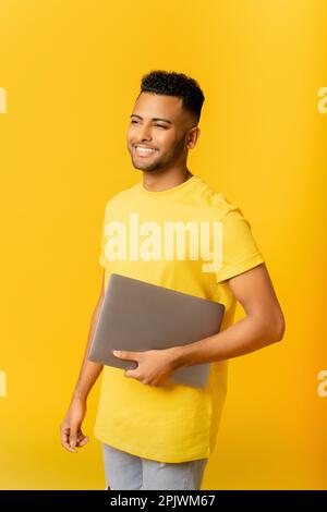 Ein gutaussehender, fröhlicher, lächelnder Indianer, der Laptop in der Hand hält, lächelt und träumig zur Seite blickt, isoliert auf gelbem Hintergrund steht, männlicher ausländischer Student, bereit zu lernen Stockfoto