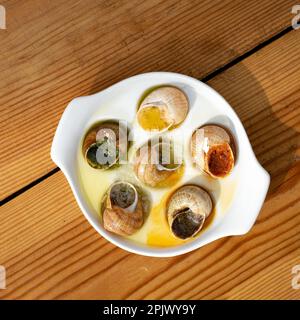 Gekochte Schnecken mit Knoblauch, Tomaten, scharfer Sauce, Kräutern, Weißwein auf einem weißen Gericht mit Holzhintergrund, französische Gourmetküche, Escargots Stockfoto