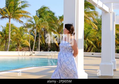 Porträt einer Frau in Sommerkleidung, die neben der weißen Säule am Swimmingpool steht und Kokospalmen im Hintergrund hat. Urlaub im tropischen Resort Stockfoto