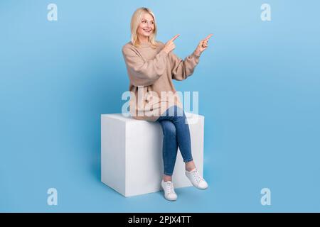 Ganzkörperporträt der positiven Großmutter Sitzen Podium sehen direkt Finger leer auf blauem Hintergrund isoliert Stockfoto