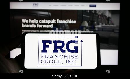 Person, die ein Mobiltelefon mit dem Logo der US-amerikanischen Franchise Group Inc. Besitzt (FRG) auf dem Bildschirm vor der Webseite des Unternehmens. Konzentrieren Sie sich auf das Display des Telefons. Stockfoto