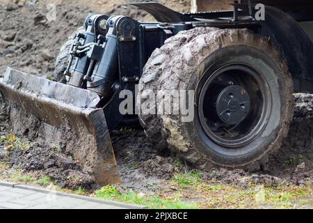 Radtraktor mit Schaufel räumt an bewölkten Regentagen den Boden frei. Schmutzige Bulldozer-Schaufel Nahaufnahme... Stockfoto