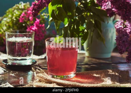 Rustikaler ästhetischer Tisch im Freien mit Glas Wasser und Kompott. Ästhetisches Picknick mit Flieder im Frühling an einem sonnigen Tag. Harte Schatten. Stockfoto