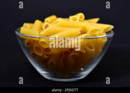 Weizenpasta in einer Glasschüssel auf schwarzem Hintergrund. Trockene Pasta in einem durchsichtigen Behälter. Stockfoto