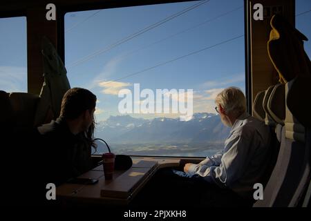 Ein alter Mann und ein Paar genießen den Blick auf die schneebedeckten Berge und den Genfer See in einem Zug von Montreux nach Gstaad mit dem Golden Pass Express Stockfoto