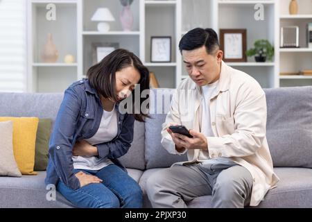 Familienpaar Mann und Frau, die zu Hause auf dem Sofa sitzen, asiatischer Familienvater, der versucht, einen Arzt für eine kranke Frau zu rufen. Stockfoto