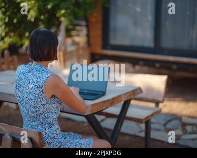 Glückliche junge asiatische Frau mit Laptop, die sich draußen in einem winzigen Haus im Sommergarten ausruht, Wochenendausflug und eine Idee für ein abgelegenes Büro, am frühen Morgen. Winzige Häuser A Stockfoto