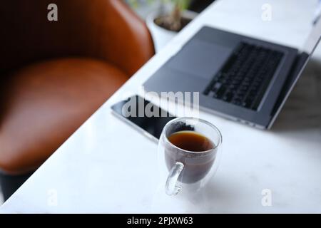 Arbeitsplatz mit Telefon-Laptop und transparenter Tasse Kaffee im Home Office auf weißem Marmorschreibtisch mit braunem Lederstuhl und grünem Baum im Hintergrund Stockfoto