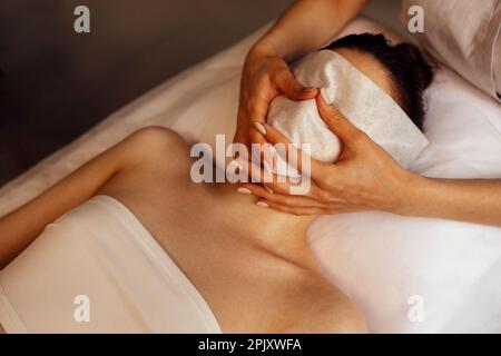 Kosmetische Massnahmen im Schönheitssalon oder in der Klinik. Das Gesicht einer jungen Mandantin ist mit einem speziellen Tuch bedeckt und die Hände einer Masseurin mit einer weißen Maniküre Stockfoto