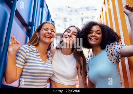 Drei süße Teenager-Mädchen in lässiger Kleidung auf bunten Versandbehältern. Weibliche Teenager verschiedener Nationalitäten in der Nähe von Metallkleckern Stockfoto